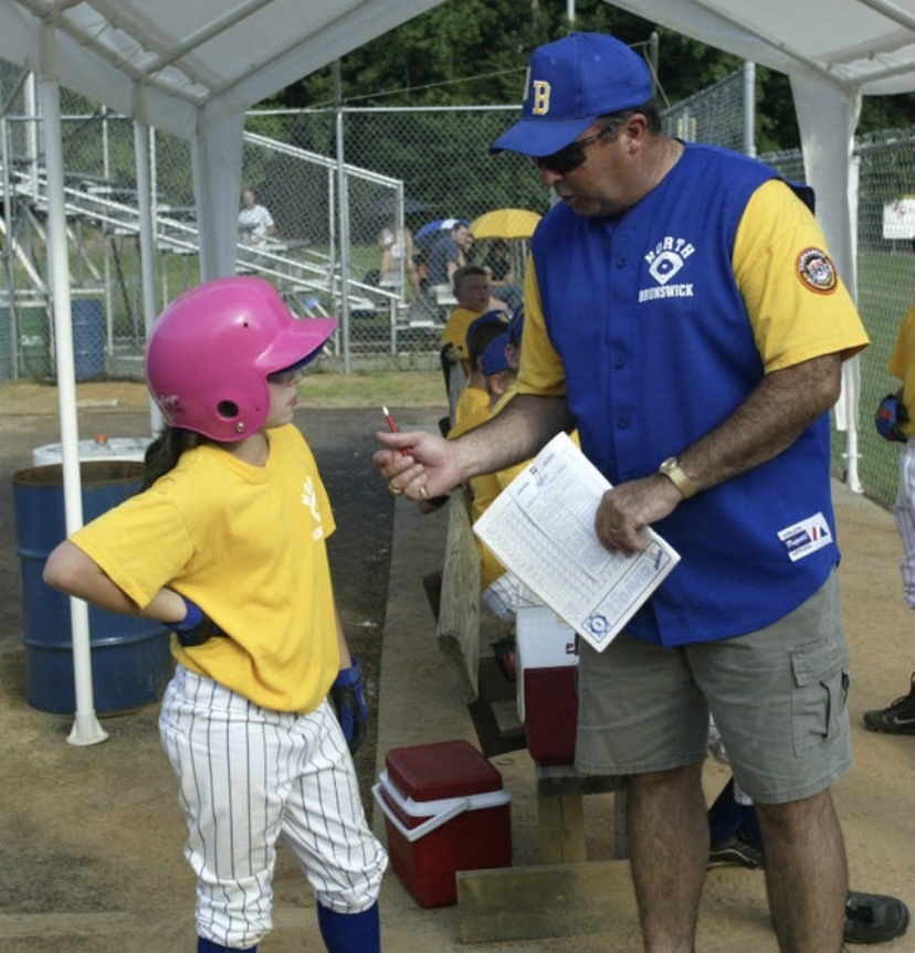 Mr. Jim Muldowney coaching Ms. Marylynn Muldowney during a baseball game.
“Some of my favorite memories growing up were the countless hours we spent at the ball field. Whether it was for my games, my brother’s, or just watching other kids play, we were always there, and my dad was always coaching. He’d usually start off standing alone by the backstop, but as the game went on, more and more people would gravitate toward him. By the end, he’d be surrounded by a group of fathers, all eager to talk baseball. What I loved most about those moments was how often people looked to him for insight. Any time a close call was made or a big play happened, coaches and fans would approach him mid-inning, asking what he thought. He was a walking encyclopedia of the game, and everyone respected his opinion.” — Ms. Marylynn Muldowney, daughter of Muldowney
